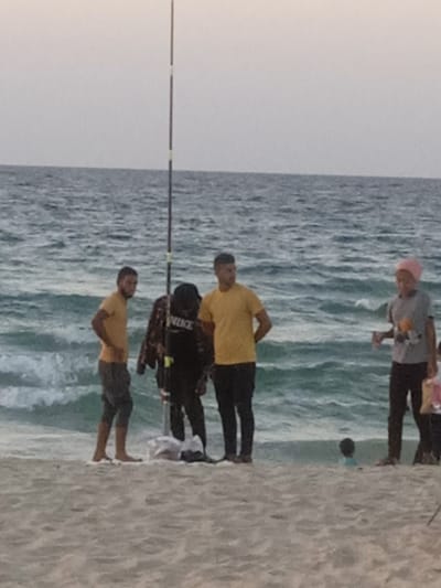 The image displays a beach scene in Al-Mawasi, Palestinian Territories. The sky is uniformly light gray, indicative of overcast conditions or twilight. The sea in the midground and background features choppy, turquoise-blue water with visible wave action. The foreground consists of light beige sand.
Six individuals are present on or near the sandy shoreline. A tall, slender fishing rod, dark with light-colored segments, stands vertically in the sand, slightly to the left of the image's center.
From left to right:
1. A male figure stands barefoot on the sand, wearing a mustard yellow short-sleeved t-shirt and dark, knee-length shorts. His hands are placed on his hips.
2. A person in a black long-sleeved shirt with a white "NIKE" logo on the back, black pants, and white shoes stands behind the fishing rod. Several white plastic bags and other items are on the sand directly in front of this individual.
3. A male figure stands on the sand, wearing a mustard yellow short-sleeved t-shirt and dark long pants. His hands are clasped behind his back.
4. A female figure stands closer to the water's edge, wearing a gray short-sleeved t-shirt, dark long pants, and a pink head covering. She holds a transparent cup or bottle in her right hand.
5. Partially obscured by the female figure, a child's lower torso and legs are visible, wearing dark pants.
6. A small child, wearing a light blue top, is partially submerged in the shallow water, with only their head and upper torso visible.