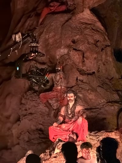 In Gollagutta, India, a scene unfolds within a rocky cavern, illuminated by warm, artificial lighting. Two men, adorned in traditional Indian attire with red fabric draped around their lower bodies and intricate necklaces, are seated. One man, in the foreground, has a prominent mustache and a white marking on his forehead, suggesting a religious or ceremonial context. He sits cross-legged. Behind him, another man, similarly dressed, holds a trident-like object. A large camera rig, mounted on a robotic arm, is positioned to the left of the frame, indicating that this scene is being filmed or photographed. The cavern walls are rough and textured, with visible rock formations and shadows. In the lower right of the image, the silhouettes of several people are visible, observing the staged scene.