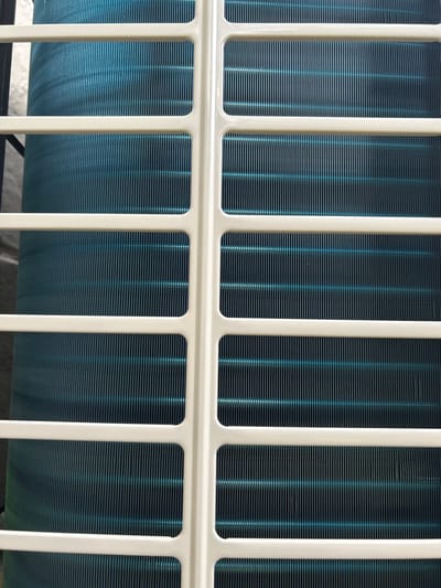 A close-up shows a white metal grid partially obscuring a blue heat exchanger. The grid consists of vertical and horizontal bars forming rectangular and square openings. Fine, horizontal lines characterize the heat exchanger, giving it a textured appearance. The lighting is diffuse, casting soft shadows within the openings. The overall composition is symmetrical and emphasizes the repetitive patterns of the grid and the heat exchanger fins.