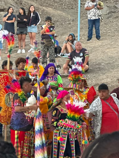 A lively group of people fills the scene, many adorned in vibrant, colorful traditional Andean clothing. Performers in feathered headdresses and intricately patterned garments mingle with onlookers. A musician plays a brass instrument on the left. Several individuals hold decorated banners or flags. The setting appears to be an outdoor event or festival.