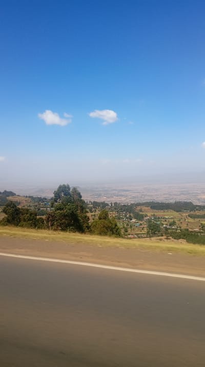 A scenic vista under a bright blue sky with scattered white clouds. A hazy, expansive valley is visible in the distance, dotted with hints of settlement. In the mid-ground, a lush, green landscape gradually slopes down towards the valley. A section of dark asphalt road with a white line bisects the foreground, suggesting the photo was taken from a moving vehicle.
