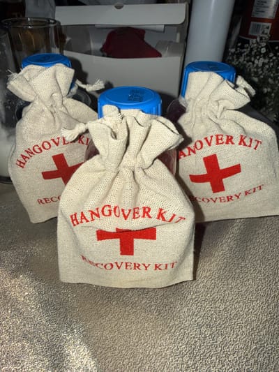 Three small drawstring bags labeled "Hangover Recovery Kit" with a red cross symbol are stacked vertically. Each bag is tied at the top and appears to contain a bottle with a blue cap. The bags are light tan or beige in color and appear to be made of burlap or a similar material. They sit on a textured surface, with other items visible in the background including a box and glass bottles.