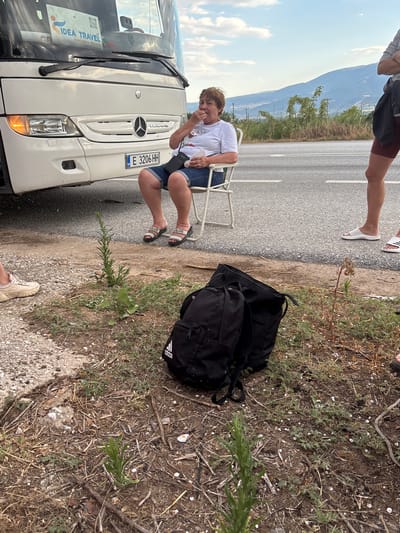 An overhead shot captures a scene on the side of a road, likely in Greece, with a white tour bus prominent in the background. A woman, appearing to be of middle age and wearing a white t-shirt and denim shorts, sits on a white folding chair, eating from a small white packet. She is positioned near the front of the bus, to the right of the driver's side. Her feet are clad in white slide sandals. A black backpack with the Adidas logo is placed on the ground in the foreground. To the right, another person's legs are visible, wearing burgundy shorts and white slide sandals, suggesting they are standing. The environment is dry and dusty, with sparse vegetation and scattered debris on the roadside, leading up to a paved road with white markings. In the distance, rolling hills under a pale blue sky provide the backdrop. The license plate of the bus reads "E 3206 HH". The overall impression is a pause or stop along a journey, with individuals taking a moment to rest and perhaps eat.