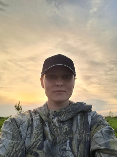 A person wearing a camouflage hooded sweatshirt and a dark baseball cap is the primary subject. The background features a sky with streaks of orange and white, suggesting either sunrise or sunset. Below the sky, a strip of green vegetation is visible. The photo appears to be taken outdoors in a natural setting; there is no indication of the location being in the city of Шешурга, Russia.