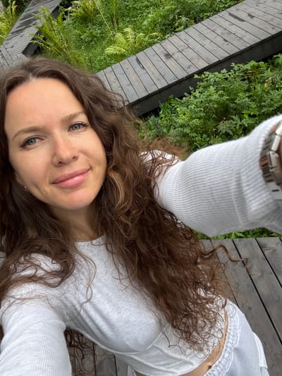 A medium shot captures a young woman with long, wavy brown hair, light blue eyes, and a light complexion. She is wearing a white long-sleeved shirt and is looking directly at the camera, smiling softly. Her left arm is extended, holding a smartphone for a selfie. The background consists of a wooden walkway and lush green vegetation, suggesting an outdoor environment. The scene is set on the outskirts of Nizhnekayancha, Russia, where wooden pathways are often integrated into natural landscapes for pedestrian access.