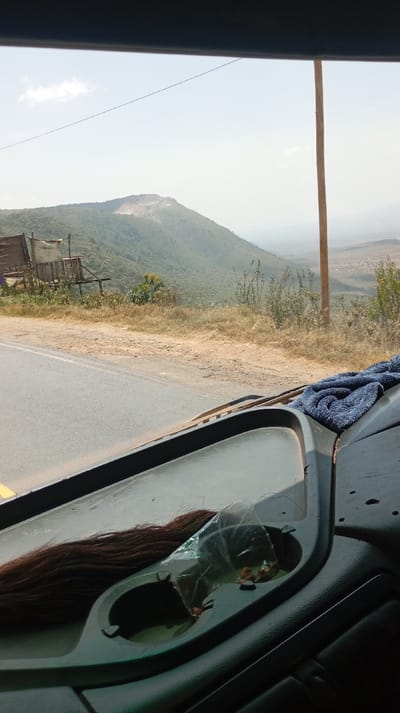 The image depicts a rural mountainous landscape in Lari/Kirenya ward, Kenya, viewed from the interior of a vehicle. An asphalt road with a dirt shoulder runs from the lower left to the mid-center. On the left side of the road, a rustic wooden structure, possibly a stall or shelter, covered with light-colored fabric, is visible above dry vegetation. A large, verdant hill with a prominent lighter-colored, excavated area (likely a quarry) near its summit dominates the left and center background. Further to the right, a broad valley with scattered vegetation and hints of distant structures extends into the background under a light blue sky with sparse clouds. A tall wooden utility pole stands vertically on the right side, and a thin power line traverses the upper sky. Inside the vehicle, on the dark dashboard, a long strand of dark brown hair rests to the left. A clear plastic bag containing small, orange-brown, oval-shaped objects is partially placed in a cup holder-like depression, and a folded blue towel is situated on the far right. No individuals are visible.