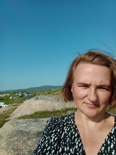This is a selfie taken outdoors with a clear blue sky. A woman with shoulder-length auburn hair is in the foreground, smiling slightly. She is wearing a black and white patterned dress. Behind her, a grassy hillside slopes down, dotted with several white camper vans and cars, suggesting a campsite. In the distance, low hills and a settlement are visible. The foreground features large, grey rocks and patches of green grass, indicative of a natural landscape. The scene appears to be in a rural area, possibly near a coast, given the open landscape and clear horizon. The provided location is Carnboy, Ireland.