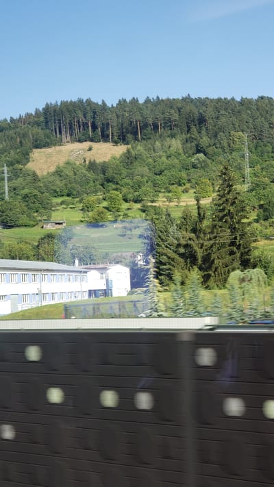 The image depicts a sunny day with a clear blue sky above a densely wooded hillside. The hillside is covered with a mix of evergreen and deciduous trees, with some patches of dry grass visible on the slope. In the mid-ground, there is a long, white, two-story building with blue-tinted windows. Adjacent to this building is another, smaller white structure with a prominent blue sign. Several tall evergreen trees are situated in front of the buildings and on the right side of the image. A power line pylon is visible on the far right of the hillside. The foreground of the image is dominated by a dark, horizontal barrier with regularly spaced, lighter-colored circular or oval shapes, suggesting it might be part of a fence or a sound barrier along a road or railway. The image appears to have been taken from a moving vehicle due to the slight motion blur. The location is identified as Bytča, Slovakia.