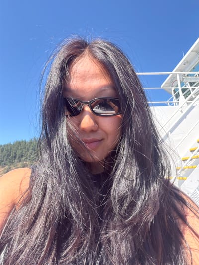 A woman wearing sunglasses and a black top is taking a selfie outdoors under a clear blue sky. She has long, dark, wavy hair that is blowing in the wind. In the background to the right, there is a white structure with railings and yellow accents, possibly part of a boat or building. To the left, there is a green, treed hillside. The overall lighting suggests it is daytime. The image is framed as a close-up of the woman's face and upper body.