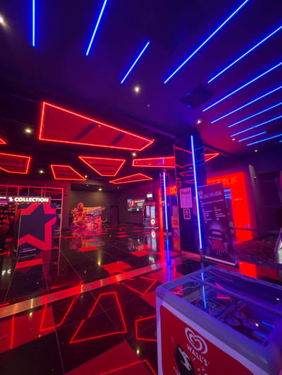 This is an interior shot of a cinema lobby in Eastbourne, UK. The walls are primarily dark and decorated with geometric shapes outlined with neon red and blue lighting. The blue lights are installed on the ceiling, aligned to create converging lines.
Signs indicating "CINEMA 1-5" and "CENS 6-8" direct patrons. There's signage advertising "MUNCHBOX" and various cinema snacks, including popcorn, sweets, and drinks. A cooler with "WALL'S" branding is visible, likely filled with ice cream. There's also a standee or display for "The Mandalorian and Grogu".
The floor is a dark, reflective surface with red geometric patterns, mimicking the lighting on the walls. The lighting, the color scheme, and signage indicate a modern, entertainment-oriented setting. There are no people explicitly visible in the scene.