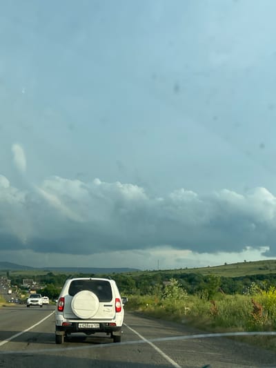 This media file shows a view from a vehicle traveling on a road. The primary subject is the rear of a white SUV with a spare tire mounted on the back. The license plate is visible and reads "K428KB 126". Following behind the white SUV are other vehicles, suggesting a procession or a busy road.
The scene is a rural or semi-rural landscape with green hills and vegetation on the left side of the road. The sky is partly cloudy with dramatic, dark clouds on the left, hinting at potential weather changes, while the right side of the sky appears lighter. The time of day appears to be daytime, given the lighting.
The setting is a road, likely in Russia, given the license plate and location context. The road itself is paved and has white lane markings. The overall impression is a journey through a scenic, possibly somewhat remote, area. There are no people clearly visible in the frame. The image is taken from a slightly elevated perspective, looking forward and down at the road and the landscape.