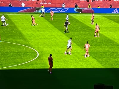 This is a high-angle, eye-level shot of a soccer match in progress. The field is green with white markings. In the foreground, a man in an orange uniform with the number 11 on his back stands facing left. In the middle ground, several players in white and orange uniforms are positioned across the field. The background shows a stadium with a red seating area and blue advertising boards with the text "F50." The image does not provide any specific information or context related to the city of Delbrück, Germany.