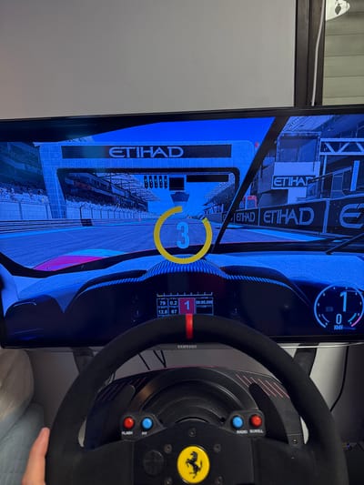A television displays a racing simulation game, showing a countdown from 3 at the starting grid of what appears to be the Abu Dhabi circuit, indicated by "ETIHAD" signs. Game data overlays include position, lap time, speed, and other statistics. In front of the screen, a black racing wheel with red accents and a yellow Ferrari logo is held. Buttons labeled "FLASH", "PIT", and "RADIO SCROLL" are visible on the wheel.