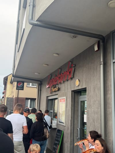 This media file captures a group of people gathered outside a pizzeria in Skawina, Poland. The scene appears to be during the daytime, with natural light suggesting it could be late morning or early afternoon. The weather seems pleasant.
The central focus of the image is the entrance to the pizzeria, adorned with prominent red and silver signage that reads "RILU'S" with "Adamus" underneath. A menu board is visible to the right of the entrance, and a Pepsi sign is also present.
Several individuals are visible in the frame. Two women in the foreground are enthusiastically eating large slices of pizza, with one woman leaning in to share a bite with another, conveying a sense of enjoyment and shared experience. Other people are seen standing and queuing, suggesting they are either waiting to order or have just received their food.
The architecture of the building features modern grey tiling on the facade, contrasting with a lighter-colored awning or roof structure. Plumbing and electrical conduits are visible on the exterior. The overall atmosphere appears casual and communal, with people enjoying a meal together on what seems to be a relatively normal day.