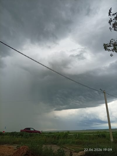 The image captures a dramatic sky with heavy, dark storm clouds dominating the upper portion. In the middle ground, a dark red sedan is parked on the side of a road, bordered by lush green vegetation. A utility pole with a light fixture stands to the right, and power lines stretch horizontally across the frame. The background shows a flat, open landscape under the brooding sky, with faint structures visible in the distance. The bottom right corner displays a timestamp: "22.06.2026 19:09". The scene appears to be an exterior shot of a rural or semi-rural area, possibly on the outskirts of Sarytogay, Kazakhstan, given the information provided.