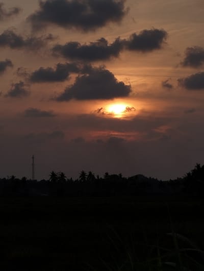 The image shows a sunset in Kutameuligoe, Indonesia. The sun is partially obscured by clouds, creating a warm, orange glow in the sky. In the foreground, there is a dark landscape with silhouettes of trees, a communications tower and some grass. The scene evokes a sense of tranquility and the beauty of nature.