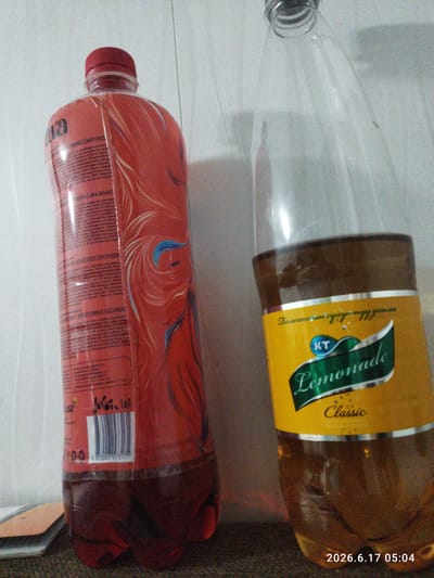 The image shows two plastic beverage bottles on a textured surface, likely a floor or table, with a white wall in the background. The bottle on the left is red and appears to contain a red liquid. It has a red cap and a label with various text and decorative patterns. A barcode is visible on the lower portion of this bottle. The bottle on the right is clear and contains a yellowish liquid, presumably lemonade, as indicated by the label. This label is predominantly yellow with a green ribbon graphic that reads "Lemonade Classic" in a stylized font. Above the green ribbon, there is text in what appears to be Cyrillic script, and a small blue circle with "KT" inside. A timestamp "2026.6.17 05:04" is visible in the bottom right corner of the image, suggesting this is a digital photograph with embedded metadata. The lighting appears to be from the front and slightly to the left, casting shadows. The scene does not provide specific geographic indicators for Bökönbaev, Kyrgyzstan.