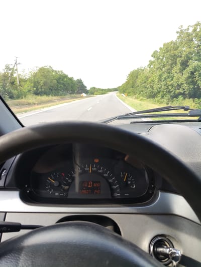 The image is a first-person view from inside a vehicle, showing the dashboard and the road ahead. The steering wheel is prominent in the foreground. The dashboard displays various gauges and indicators, including a speedometer showing a speed of 110 km/h, an odometer reading 628607 km, and a clock showing 16:50. A digital display also shows "240" which could be related to engine hours or another metric. The road ahead is asphalt and appears to be a rural or suburban highway, flanked by trees and vegetation. The sky is overcast or bright white, obscuring any details of the sun. The location is identified as Podgoreni, Moldova.