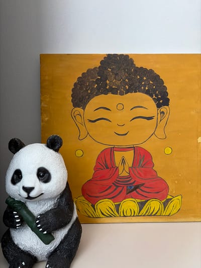 A panda figurine and a painting of Buddha are positioned on a white surface, with a gray wall in the background. The panda, which is black and white, is holding a green bamboo stalk in its paws. The painting depicts a stylized, cartoon-like Buddha with closed eyes, wearing a red robe and sitting on a yellow lotus flower. The background of the painting is a vibrant golden yellow. The scene is well-lit, with the light source appearing to be from the front and slightly to the left, casting soft shadows. The overall impression is one of peaceful decoration, possibly in a home or a shop. The location in Kyzyl, Russia is not discernible from the visual information provided in the image.