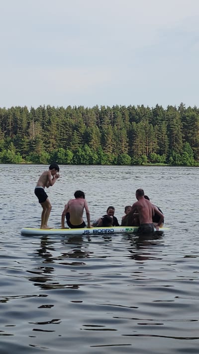 Six individuals are gathered on and around a stand-up paddleboard in a body of water, with a densely wooded shoreline in the background under a lightly clouded sky. One person stands on the paddleboard, facing away from the camera and appears to be mid-motion, possibly gesturing or looking down. The other five people are seated or partially submerged around the paddleboard. The paddleboard is white with yellow accents and features the text "JS BOARD" and a "CE" marking. The water is rippled, reflecting the sky and the surrounding trees. The environment suggests an outdoor recreational setting, likely a lake or calm river, typical of areas near forests. The location is identified as Nutromo, Russia.