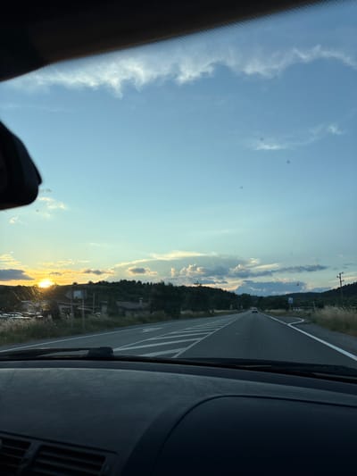 The image is a photograph taken from inside a vehicle, looking out the windshield at a road and the surrounding landscape. The photo is oriented vertically, with the sky on the left and the road on the right.
The subjects include the road, a few distant cars, trees, a utility pole, and a bright sunset. The scene is a rural or suburban area, possibly a highway, as indicated by the clear lane markings. The setting appears to be Blatino, Bulgaria, based on the provided context.
The activity taking place is a drive. The time of day is sunset, evidenced by the low angle of the sun and the warm, golden light illuminating the clouds and landscape. The weather appears clear with scattered clouds.
Notable details include the presence of white lane markings on the asphalt road, creating a leading line towards the horizon. The trees lining the road are lush and green, suggesting a temperate climate. The sky transitions from a bright blue to a softer yellow and orange near the horizon where the sun is setting. The interior of the car, including part of the dashboard and windshield wipers, is visible in the foreground, adding to the perspective. There is no visible text in the image.
