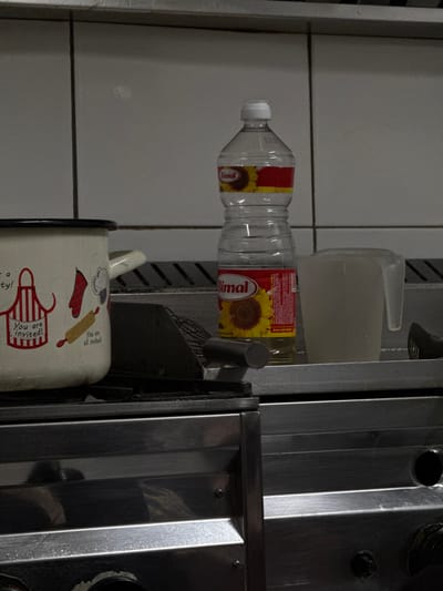 The scene is an interior view of a kitchen or food preparation area in Brčko, Bosnia and Herzegovina. No individuals are present, and no active actions are depicted. The background consists of a white tiled wall with horizontal grout lines, above a metallic work surface. A white enamel pot with a black rim and a white handle is positioned on a black grate, which is part of a stainless steel cooking range located at the bottom of the frame. The pot is decorated with red and black illustrations, including an apron, oven mitt, and rolling pin, accompanied by the English phrases "You are invited!" and "You are all invited!". To the right of the pot, a clear plastic bottle of Bimal brand sunflower oil, featuring a white cap and red and yellow labels with sunflower imagery, stands on the metallic surface. Further to the right, a translucent white plastic measuring jug partially filled with a light-colored liquid is visible. Various kitchen utensils, including a dark wire whisk, a silver cylindrical object, and dark tongs, are partially observable on the metallic surface between the pot and the jug. The lower section of the image shows the brushed stainless steel face of the cooking range, including indistinct circular control elements.