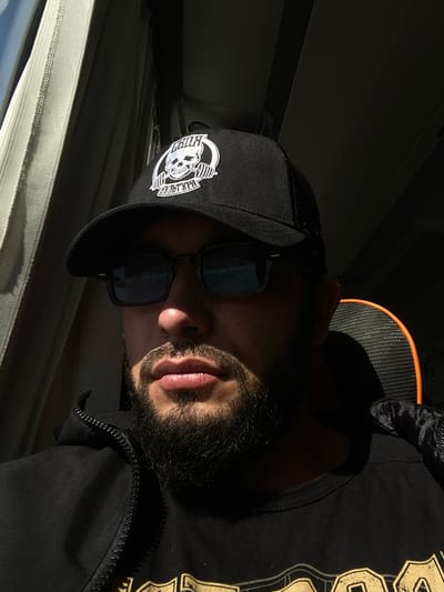A man wearing a black baseball cap with a skull logo and sunglasses is captured in a close-up shot, seated indoors, likely on public transport with a window to his left. The sunlight streams in, creating strong shadows across his face. He has a beard and is wearing a black hooded jacket zipped up, revealing a black t-shirt underneath. The background includes a window with light-colored curtains and a dark ceiling. The man appears to be in Vitino, Russia, based on the provided context.
