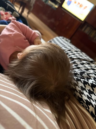 An overhead, slightly angled photograph captures a young child resting indoors, likely in a living room, in Krapivka, Russia. The child, visible from the top of their head and upper back, has light brown hair and is wearing a long-sleeved pink top. They are positioned on or next to a striped surface, which is light brown or beige with prominent white stripes. Behind the child, a black and white fabric featuring a houndstooth pattern is visible, suggesting a blanket or pillow.
In the blurred background to the left, a brightly lit rectangular screen, presumably a television or tablet, displays colorful imagery that appears to be a cartoon or show. Dark wooden furniture or paneling can be seen further back. The child's posture, with a bent arm, suggests a state of relaxation, possibly while watching the screen or simply resting. The scene conveys a calm, domestic atmosphere. No visible text is present in the image. The time of day and external weather conditions are not discernible from the indoor setting.
