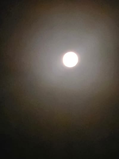 The image depicts a bright, circular light source, likely a lightbulb, positioned centrally in the frame. The light emanates outwards, creating a halo effect against a dark, textured background that appears to be a ceiling. The halo is somewhat diffused, suggesting a slight haze or dust in the air. There are subtle variations in the background's tone, transitioning from a lighter gray around the light source to a darker, almost black hue at the edges of the image. The overall impression is one of an indoor environment, possibly a room or an open space, illuminated by a single overhead light. The image does not contain any people, animals, or specific details that would pinpoint its location within Beer Tuvia Regional Council, Israel, or any other specific urban or rural setting.