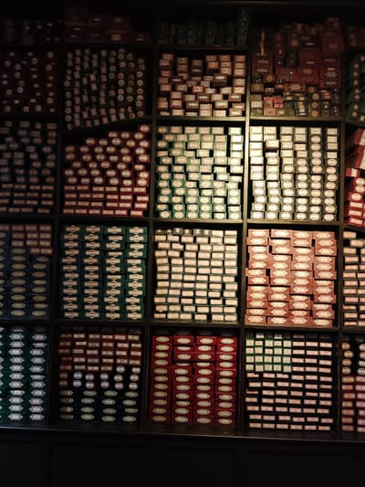 The image depicts a wall of wand boxes at Ollivanders Wand Shop in the Warner Bros. Studio Tour London, located in Watford, United Kingdom. The boxes are arranged in a grid-like structure, filling the shelves from floor to ceiling. The boxes are of various colors and designs, including green, red, brown, and black, with different shapes and labels on each box. There are no people visible in the shot.