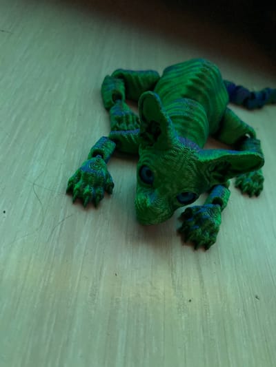 An articulated, 3D-printed toy, resembling a stylized creature like a dragon or a Sphynx cat, is positioned on a light-colored, possibly wooden or laminate, surface. The toy is predominantly green with blue accents, featuring a textured, layered appearance characteristic of 3D printing. It has a distinctive head with large, pointed ears and dark, recessed eyes. Four limbs with clawed feet extend from its body, and a long, segmented tail is partially visible stretching behind it. The creature appears to be lying down, with its head slightly raised.
The surface beneath the toy has a subtle grain pattern and appears to be a floor or tabletop. There are some minor specks of dust and what appear to be thin strands of hair on the surface, particularly noticeable around the toy's front left paw. The lighting is diffused, casting soft shadows and highlighting the texture of the 3D print. No specific activities are taking place, as the toy is stationary. The setting is indoors, but no further details about the room or location are visible beyond the surface and the toy itself. There is no visible text in the image.