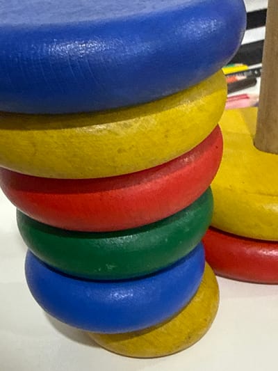 The media file shows a close-up, side-view of a stack of colorful, circular wooden beads, likely from a child's toy or educational material. The beads are arranged in a diagonal line from the bottom left to the top right of the frame. From left to right, the visible beads are blue, yellow, red, green, blue, and yellow. The surface of the beads appears slightly worn, with some visible texture and minor imperfections, suggesting they have been used. In the background, out of focus, are some vertical stripes of black and white, possibly part of another toy or a patterned surface. The lighting is even and appears to be natural, casting soft shadows. The overall impression is a simple, bright, and cheerful arrangement of children's play items. There is no visible text in the image.