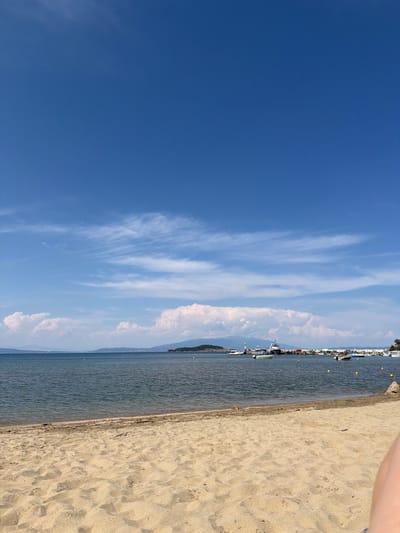 The image depicts a serene beach scene in Olympiada, Greece, on a clear, sunny day. The foreground is dominated by a sandy beach with soft, undulating textures. The sand appears light brown and is strewn with small pebbles and debris.
The middle ground features the calm, blue-green waters of the sea. The water's surface is mostly smooth, with subtle ripples indicating a gentle breeze. A few yellow buoys are visible in the water, marking a boundary or perhaps a swimming area.
In the background, a coastline stretches across the horizon, lined with distant hills and mountains. A small harbor is visible, with several boats and yachts anchored. The boats are of various sizes, including a larger vessel with a dark hull and white superstructure, and several smaller motorboats. The harbor area also shows some structures along the shoreline, possibly buildings or piers.
The sky above is a vibrant, cloudless blue, with thin, wispy clouds scattered in the distance, adding a touch of texture to the expansive sky. The overall impression is one of a peaceful, idyllic seaside location.