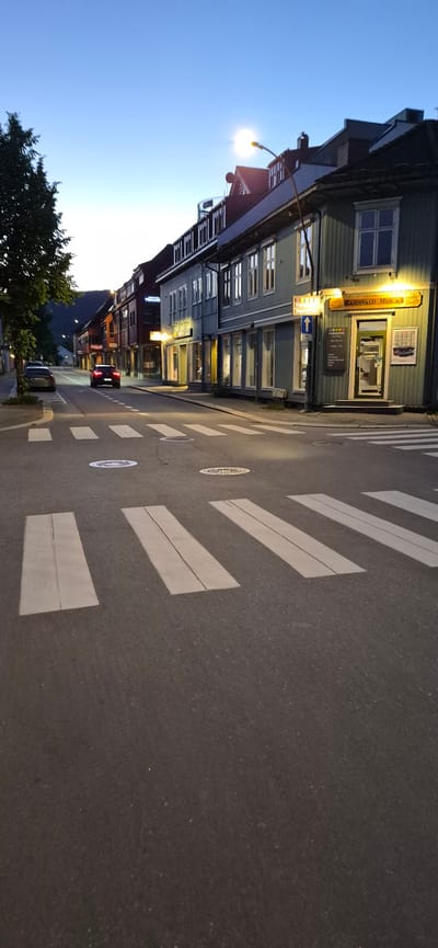 The image depicts a street scene in Lillehammer, Norway, at dusk or early morning. The foreground shows a pedestrian crossing with white stripes on asphalt. Two manhole covers with circular patterns are visible on the road.

A street with buildings lining its right side extends into the background. The buildings are of traditional Norwegian architecture, with wooden facades painted in various colors, including green and red. Some buildings have multiple stories with visible windows and pitched roofs. A street lamp with a bright light is visible in the upper right portion of the frame, illuminating parts of the buildings.

A red car is driving away from the camera on the left side of the street. Another parked car is visible further down the street on the left. The sky is a clear, pale blue, transitioning to a lighter hue near the horizon, suggesting twilight. A tree with dark green foliage is present on the far left of the image. The street appears to be in a commercial or residential area. One storefront displays a sign with "RAGNVALD MOE A/S" and what appears to be a business name in Norwegian.