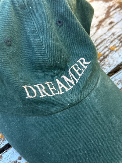 The image shows a close-up view of a dark green baseball cap. The word "DREAMER" is embroidered in white on the side of the cap, with the letters curved to follow the shape of the cap. The fabric of the cap has a textured weave. The cap is resting on a surface that appears to be weathered wooden planks, with some lighter colored paint or stain and yellow splotches visible. The lighting suggests it is daytime, with shadows indicating the light source is coming from the side. The overall setting appears to be outdoors, possibly in a rustic or casual environment, with no specific identifiable landmarks within the frame. The image is a static, close-up shot of the cap, with no activity or event depicted beyond the object itself.
