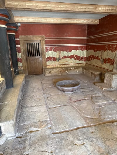 The image depicts the interior of a room with ancient architectural features in the Palace of Knossos, Heraklion, Greece. The walls are painted with a red background and adorned with horizontal white stripes and a fresco depicting griffins or similar mythical creatures amidst stylized plants. The floor is paved with large, uneven stone slabs. In the center of the room, a large, shallow stone basin rests on the floor. To the left, imposing dark columns with red and blue bands rise towards wooden beams supporting the ceiling. A wooden door with vertical slats is visible on the back wall. Stone benches are integrated into the walls, and a stone altar-like structure stands to the right. The lighting is natural, suggesting an outdoor or well-lit interior space. There are no people present in the image.