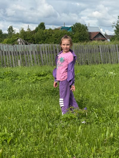 The image shows a young girl lying in a grassy field in what appears to be a rural setting. She is wearing a pink t-shirt with a graphic design and purple sweatpants. Her hair is blonde and pulled back with a purple tie. The girl is smiling and looking at the camera.
Behind the girl, a weathered wooden fence runs horizontally across the frame. Beyond the fence, trees and a few buildings are visible, including a house with a red roof and a blue building. The sky is cloudy, suggesting it may be an overcast day. The grass is lush and green, with some wildflowers scattered throughout.
The scene conveys a sense of childlike innocence and simple outdoor enjoyment. The location appears to be a village or countryside area, given the presence of the fence, rural buildings, and natural landscape. There is no visible text in the image.