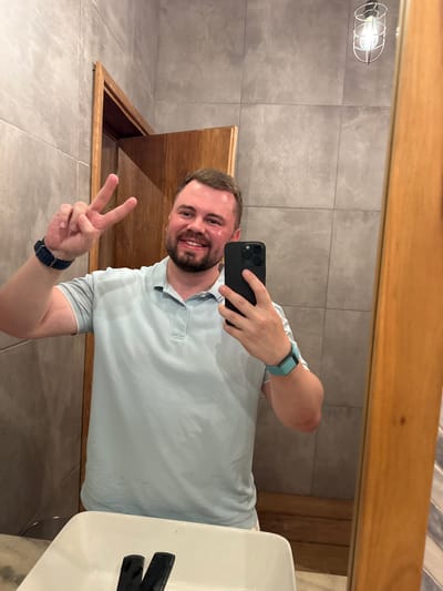 A smiling man takes a mirror selfie in a bathroom with grey tiled walls. He holds a black smartphone with a triple-lens camera. He's wearing a light blue polo shirt, a blue watch band on one wrist, and a teal fitness tracker on the other. He flashes a peace sign. A wooden-framed mirror surrounds him. A light fixture and wooden cabinet are also visible. A white sink is partially in view on the right.