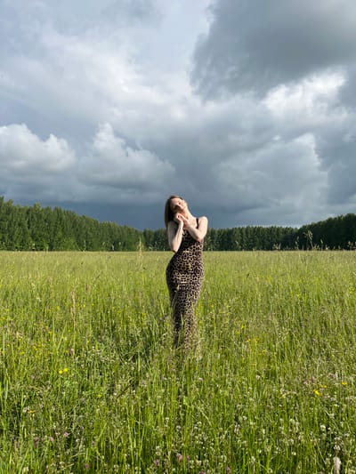 A full-body shot of a young woman in a leopard-print maxi dress stands in a field of tall green grass and wildflowers. The woman is positioned in the center of the frame, with her head tilted back and eyes closed, her hands clasped together near her face in a pose of serene contemplation. The field is lush with a variety of green grasses and small white and pink flowers, with a few yellow wildflowers interspersed. In the background, a dense line of dark green trees stretches across the horizon, forming a stark contrast against a dramatic sky filled with heavy, dark gray clouds, suggesting an impending storm. The overall lighting is subdued due to the cloud cover. The location is the outskirts of Большой Гондыр, Russia.