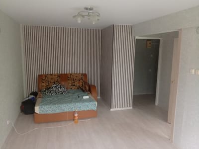 The image shows an interior view of a room, possibly a living room or a studio apartment, in Borodino, Russia. The room has light-colored walls and a light wood-grain laminate floor.
Dominating the left side of the image is a sofa with an orange-brown frame and a floral-patterned headboard. The sofa is covered with a light blue and green patterned blanket, and a black and white striped blanket is draped over it. To the left of the sofa, there are several bags, including a black backpack and a dark blue bag.
The walls are wallpapered with a vertical stripe pattern in shades of brown and off-white. A portion of the wall to the right features a similar striped wallpaper, creating a visual division.
A doorway is visible in the background, leading to another room or hallway. The doorframe is a light wood color, and the door itself is dark. A light fixture with two glass shades is mounted on the ceiling.
A bottle of orange liquid, possibly a beverage, stands on the floor near the sofa. An electrical cord runs across the floor from the wall near the sofa. The overall impression is of a sparsely furnished and possibly undecorated space.