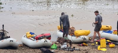 The image captures a riverside scene in Kikagati, Uganda, featuring multiple inflatable rafts and equipment on a pebbly bank adjacent to murky brown water. In the foreground, a Black male, dressed in a grey long-sleeved shirt and dark shorts, stands with his lower legs in the water, bending over a white inflatable raft loaded with yellow and brown dry bags. To his right, a White male, barefoot and wearing a grey short-sleeved shirt and dark shorts, stands on the shore holding a coiled black object. A third person's arm is partially visible on the far right.

On the pebbly shore between and around the men are various items including yellow paddles, yellow dry boxes, a yellow helmet, a white bucket filled with netting, a green plastic bag, and metal poles. Four white inflatable rafts are visible along the water's edge, three of which are laden with red, yellow, and blue dry bags or containers, and other gear. The water in the background is brown and shows scattered green aquatic vegetation.