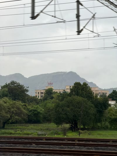 The image captures a view from a moving train in Saphale, India, with overhead railway electrification lines dominating the upper portion. Below the lines, a cluster of buildings, including a light-colored apartment complex with the name "KURLA COMPLEX" visible in red lettering on its rooftop, is nestled amongst lush green trees. In the background, rolling hills shrouded in mist or clouds suggest a slightly overcast day. The foreground features train tracks, indicating the perspective of someone traveling by rail. The environment appears to be a mix of urban development and natural landscape, characteristic of a semi-urban or suburban area.