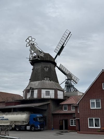 In Scholen, Germany, a large, traditional windmill stands prominently, its wooden sails angled against a cloudy sky. The windmill is integrated into a complex of brick buildings. To the left of the windmill, a blue tanker truck with the words "Futter Landhandel" emblazoned on its side is parked under a covered area. To the right of the windmill, a red brick building with white-framed windows is visible. The scene appears to be a commercial or industrial site, possibly a feed mill or grain processing facility, given the presence of the windmill and the truck. The ground is paved, and the overall atmosphere is industrial and overcast.
