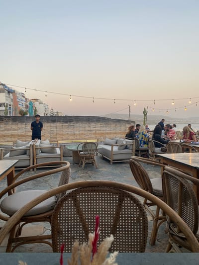 The image depicts an outdoor dining area with a view of the city of Taghazout, Morocco. In the foreground, several rattan chairs with light-colored cushions are arranged around stone-tiled flooring. A table is partially visible. In the mid-ground, there are more seating arrangements, including a sectional sofa with cushions, a small wooden table, and a wicker chair. A man in a dark blue polo shirt and dark pants stands behind the sofa, facing slightly to the right. Beyond the seating area, a wooden wall is visible, and behind that, buildings with white and blue facades are stacked on a hillside. A statue of a woman in blue is positioned on the hillside. String lights with Edison bulbs hang across the scene. In the background, a body of water is visible, with distant hills and a hazy sky suggesting dusk or dawn. Several groups of people are seated at tables in the background, dining and socializing. One person is taking a photo with a phone. The overall atmosphere suggests a relaxed, coastal evening setting at a restaurant or cafe.