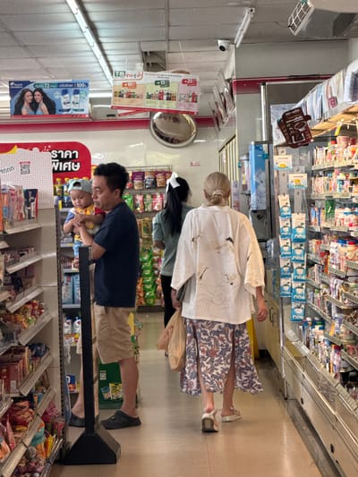 In a convenience store in Thap Sakae, Thailand, a man in a dark t-shirt and light brown shorts holds a young child wearing a blue cap and a yellow shirt. The child is held in the man's left arm. To their right, a woman with blonde hair tied back in a bun wears a white kimono-style garment with a crane design and a patterned skirt. She is carrying a plastic bag. The store shelves are stocked with various products, including bottled drinks and packaged snacks. Overhead, fluorescent lights illuminate the aisle, and a security camera is visible. Promotional signage hangs from the ceiling, displaying product information and prices. The floor is tiled.