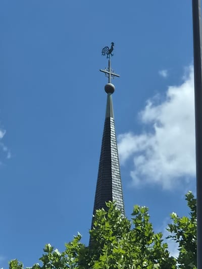 The image shows the top of a steeple with a weather vane in the shape of a rooster. The steeple is covered in dark gray slate tiles arranged in a shingle pattern. At the very top of the steeple is a cross with a globe at the intersection. The rooster is silhouetted against a bright blue sky with scattered white clouds. In the foreground, the green leaves of trees are visible, partially obscuring the lower part of the steeple. The overall impression is of a clear, sunny day. This steeple is located in Schengen, Luxembourg.
