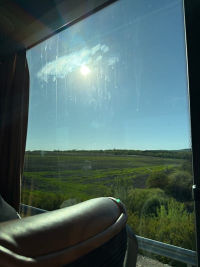 The image depicts a view through a dirty window, likely from inside a bus or vehicle. Outside, there is a field or landscape with green vegetation. A portion of a seat is visible in the foreground. The location, Malko Tranovo, Bulgaria, cannot be confirmed from the visual data alone. No people are visible. The sun is shining through the window creating glare.