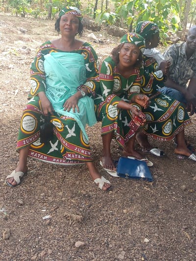 The image shows two women seated outdoors in Ninzo South, Nigeria. The women are wearing matching green dresses and head coverings, adorned with white doves and text that includes "WE SERVE THE LORD CHRIST COL 3:24" and "ZUMUNTAR MATAN ECWA". The woman on the left is wearing a light blue shawl or wrap. Both are wearing sandals. The woman on the right is holding a mobile phone. A blue handbag is visible on the ground near her feet. Behind them, a few other people are seated, including a man in a patterned shirt. The ground is covered with small rocks and debris, suggesting an outdoor, possibly shaded, area with trees.