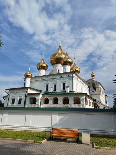 The image depicts the Assumption Cathedral in Faliukovo, Russia, under a blue sky with scattered clouds. The cathedral features multiple golden domes topped with crosses, and its white walls are adorned with ornate architectural details. In the foreground, a white brick wall encloses the cathedral grounds, with a wooden bench and a trash receptacle placed on a paved walkway adjacent to a grassy area. The overall scene conveys a sense of historical and religious significance.