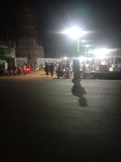 This image captures a nighttime scene on a paved road in Tundun Wada South, Nigeria. The lighting is dominated by bright streetlights, some of which have a green hue, creating a somewhat surreal atmosphere. In the background, a large building under construction, possibly a mosque or cultural center, is visible, with scaffolding surrounding it. A diverse group of people are present on the side of the road and on motorcycles, suggesting a public gathering or marketplace. Some individuals are walking, others appear to be conversing, and a few are on or near motorcycles. The road surface shows some wear and tear, with scattered debris. The overall impression is one of a lively, though somewhat dimly lit, urban environment at night.